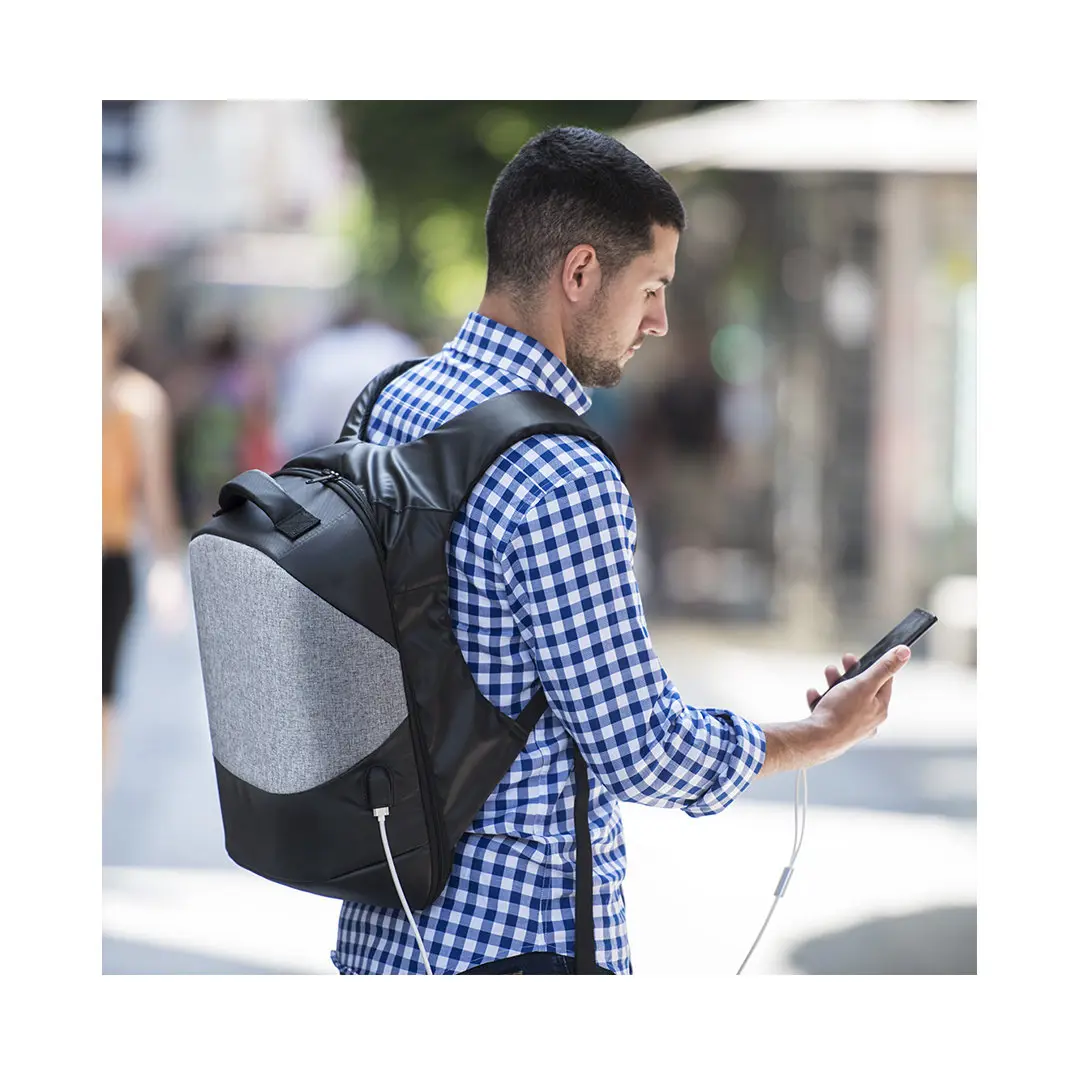 hombre de espaldas con camisa de cuadros y mochila urbana con cable USB conectado al móvil en un entorno urbano