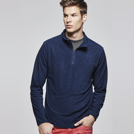 Hombre joven con sudadera polar azul de media cremallera y pantalones rojos sobre fondo gris claro