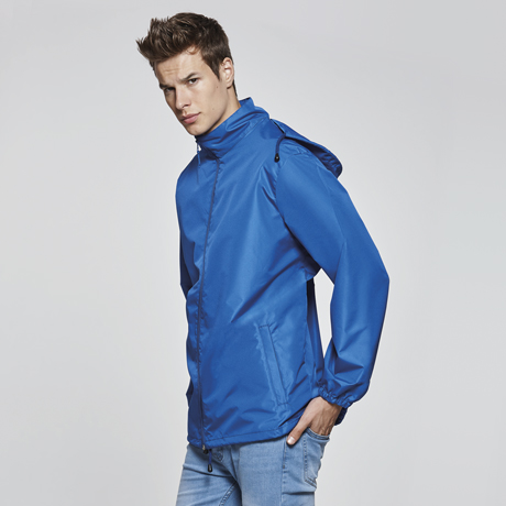 Hombre joven con cazadora azul de estilo deportivo y capucha, posando de perfil sobre fondo neutro.