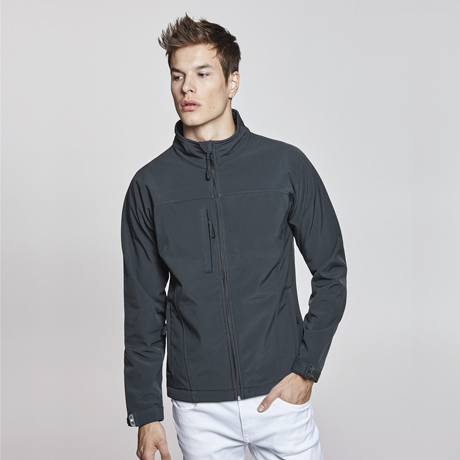 Hombre joven con chaqueta negra de cremallera y pantalones blancos sobre fondo neutro.