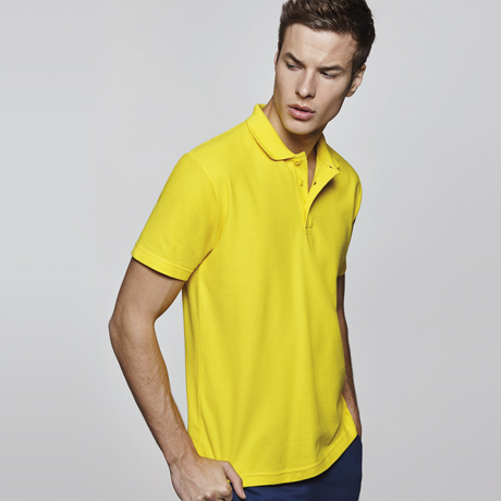 Hombre joven con polo amarillo de manga corta posando de perfil sobre fondo gris claro.