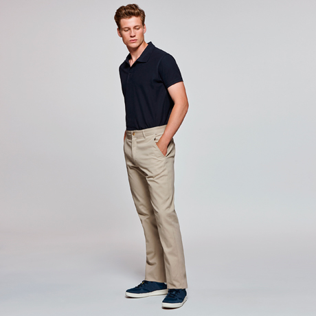 Hombre joven con polo azul marino, pantalones chinos beige y zapatillas deportivas azules sobre fondo neutro.