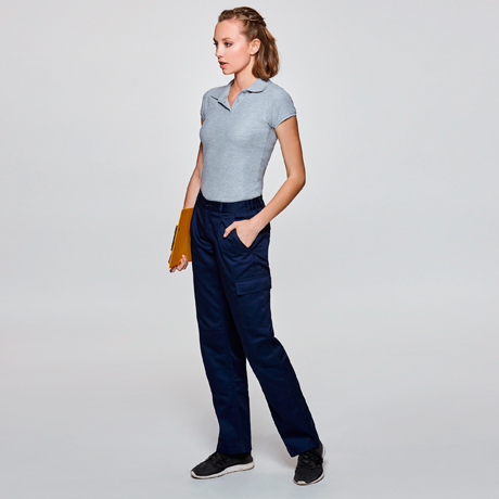 mujer joven de pie con polo gris, pantalones tipo cargo azul marino y carpeta amarilla sobre fondo neutro
