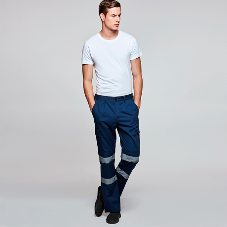 Hombre con camiseta blanca y pantalones de trabajo azul marino con bandas reflectantes sobre fondo neutro